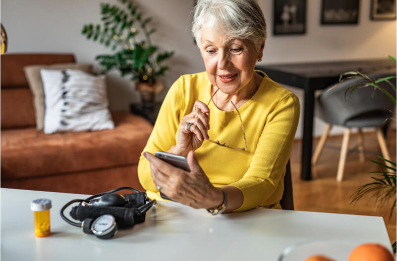 Older woman looking at her cellphone with a bottle of pills and a blood pressure monitor on the table in front of her. 