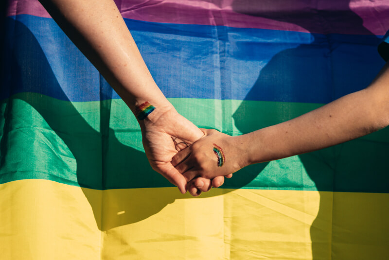 Hands held in unity against the backdrop of an LGBTQ+ flag.