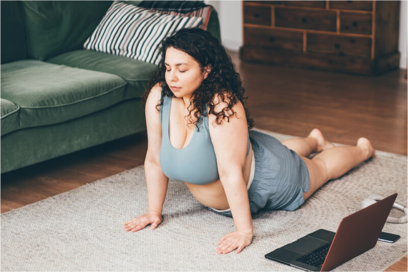 Woman with obesity wearing workout clothes and stretching on the carpet at home.