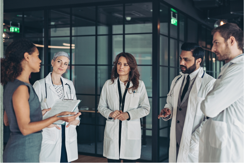 A group of doctors discussing value-based healthcare with a woman dressed formally and holding a clipboard. 